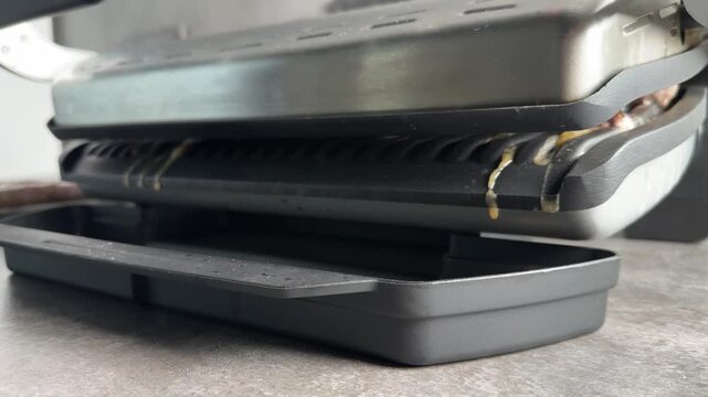 Fat and juices dripping down from hot plates of an electric contact grill into a black plastic drip tray while cooking meat products on a kitchen countertop