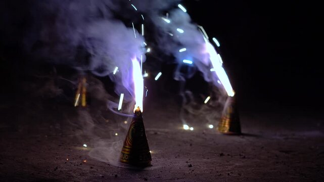 Close-up of a person using a sparkler to light an anar fountain firework during Diwali celebrations at night, with glowing flames and smoke.