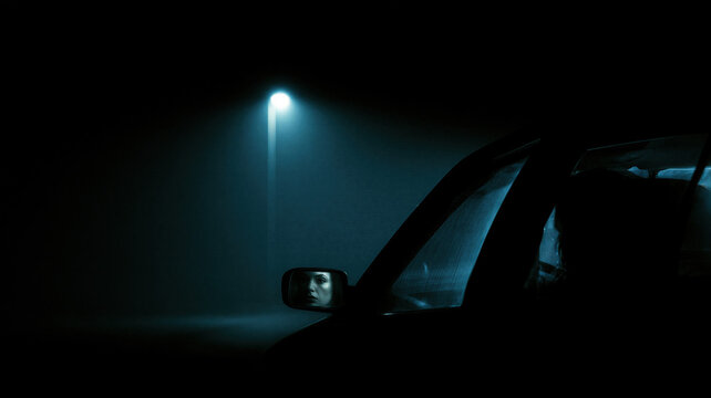 Woman looking in a rearview mirror of a car parked at night under a street light. Dark horror atmosphere with cinematic gloom and mystery. Concept of thriller scene for Halloween night.