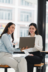 Obraz premium Two smiling Asian women collaborating using a laptop while sitting in a modern workspace