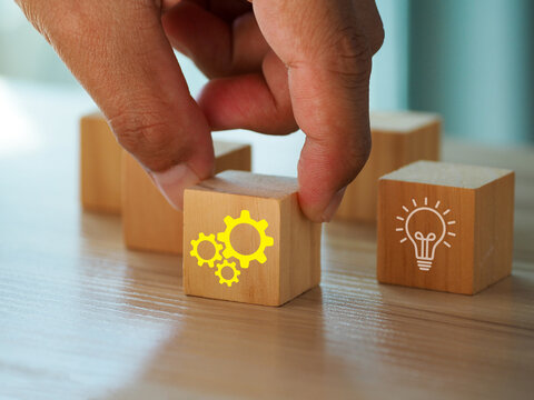 Wooden block with yellow gear icon held by hand among blocks including light bulb symbol, suggesting innovation and teamwork in business strategy and problem solving
