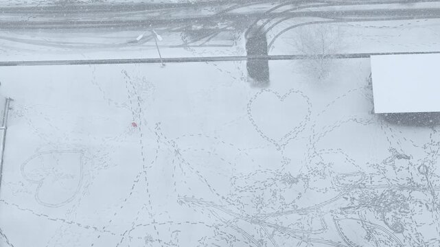 Aerial top-down view of a little girl in a red jacket walking and playing among heart-shaped footprints in deep snow during a blizzard.