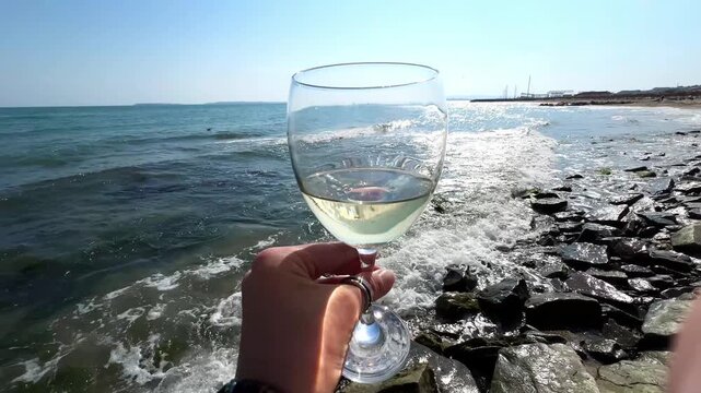 Close view of female hand holding white wine glass near rocky shoreline with sparkling sea waves and bright summer atmosphere. High quality 4k footage