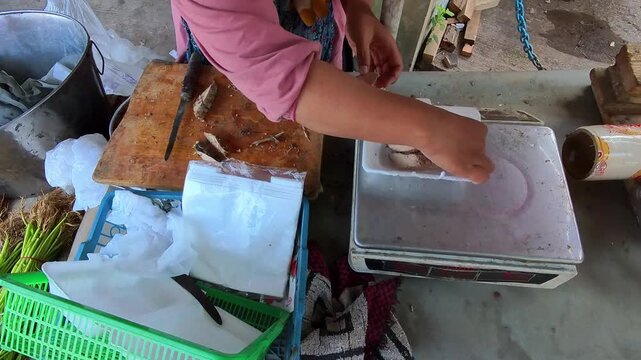 Video of Hands weighing small fish on a digital scale. Vendor prepares fish for sale at a street stall. Cutting board and plastic bags sit nearby. Fish appears fresh and ready for customers. 4K 