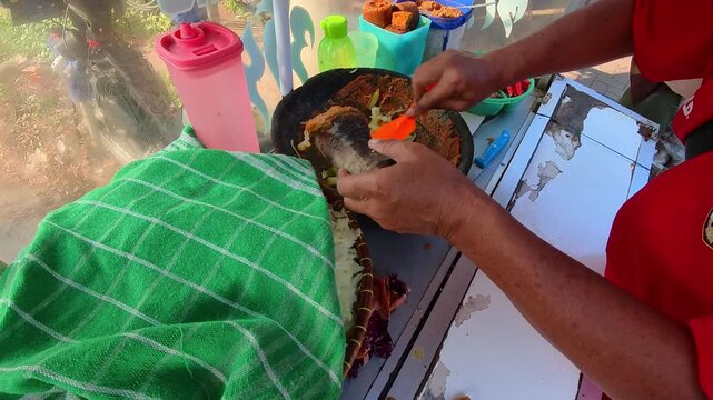 Video of Hands Mixing vegetables with peanut sauce in a skillset. Green checkered towel rests nearby. Vendor wears a red shirt while cooking. Sunlight streams through a window behind the stall