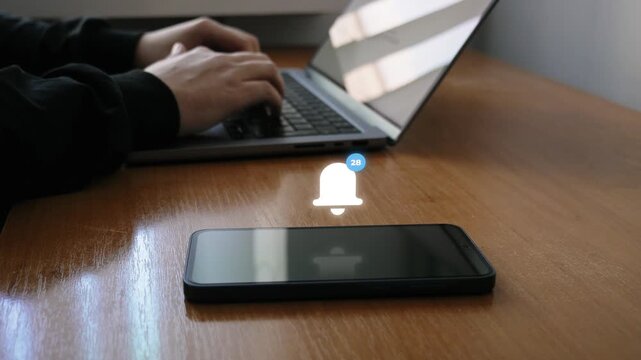 A person types on a laptop while a smartphone on a wooden desk receives increasing notification alerts, with the phone in the foreground and the laptop in the background.