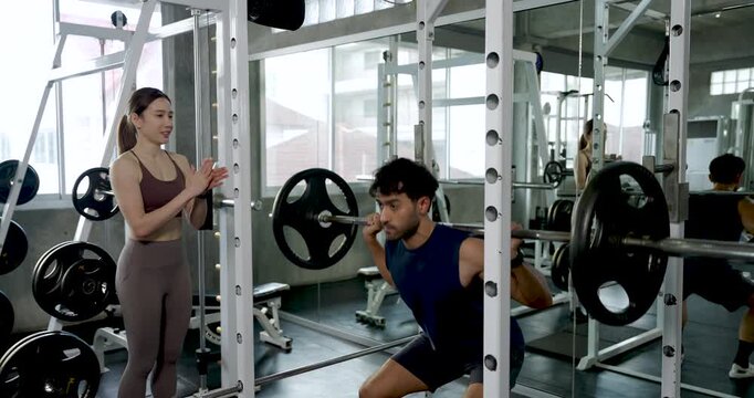 Young Asian couple taking turns with barbell squat in modern gym, man performs rack exercise while woman stands nearby guiding form, partner training, support, teamwork, motivation, wellness.