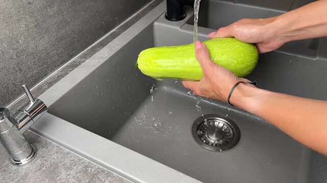 Woman's hands washing a fresh organic green zucchini under running tap water in a grey composite kitchen sink, preparing fresh ingredients for cooking