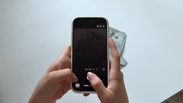 Female hands use a smartphone to photograph a stack of 100 dollar banknotes on a white background. A finance themed scene representing cash documentation, banking, payments and personal wealth.