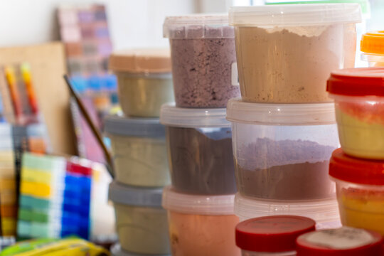 Plastic containers filled with colorful glass enamel powders and artist pigments stand on a workbench in a creative workshop, close up view with selective focus.