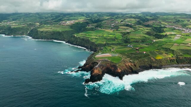 Aerial view of rugged Atlantic coastline with volcanic cliffs, ocean waves and small lighthouse on Sao Miguel Island, Azores. Green agricultural fields extend along coastal headland above ocean