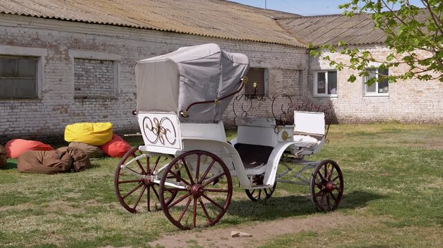 An antique white horse carriage with red wheels stands on a grassy yard beside an old brick building. Concept of historical transport, vintage romance, and countryside aesthetics.