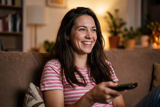 Cheerful young woman watching TV with remote control on sofa