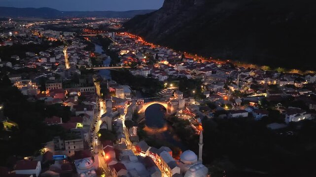 Mostar, Bosnia and Herzegovina - June 7, 2026: Drone flying left around the illuminated Old Bridge from the opposite riverbank