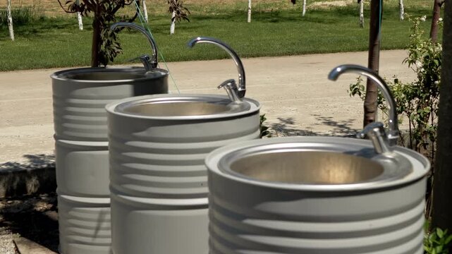 Three grey metal barrels repurposed as modern washbasins stand alongside a sunny park path. Concept of eco friendly upcycling, outdoor recreation, and creative summer glamping amenities.