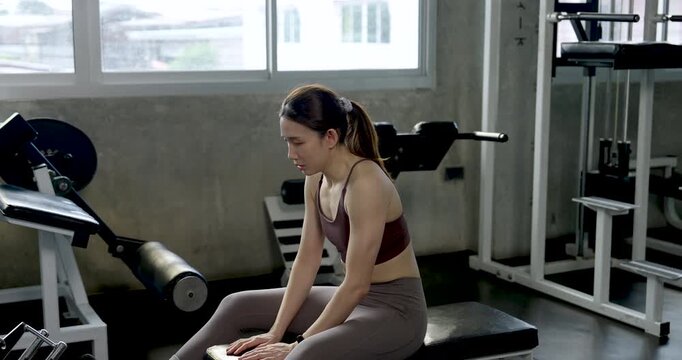 Young Asian woman rests on gym bench after seated row workout, leaning forward and catching breath between sets, solo fitness recovery moment, arm strength routine, wellness, healthy lifestyle.