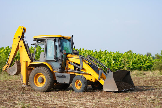 A yellow backhoe loader is digging and leveling soil in a rural agricultural field