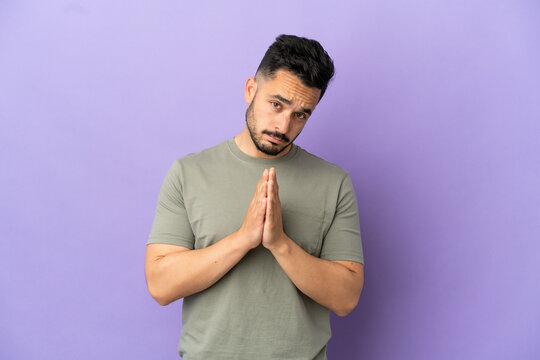 Young caucasian man isolated on purple background keeps palm together. Person asks for something