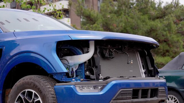 Modern blue pickup truck with significant front-end damage sitting in a car lot, its missing grille exposing the radiator and engine components while awaiting collision repair or salvage