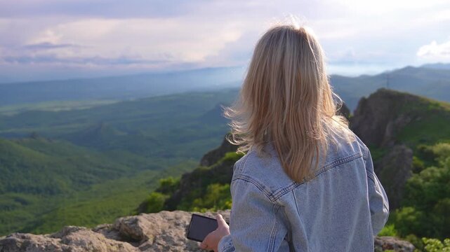Back view of a young blonde woman in a denim jacket sitting on a cliff and looking at the epic mountain landscape as her hair blows in the wind during a scenic, cloudy sunset