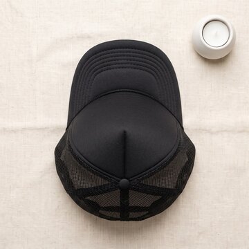 Blank black trucker hat mockup laid flat on a neutral beige fabric background indoors next to a small white candle, apparel design concept