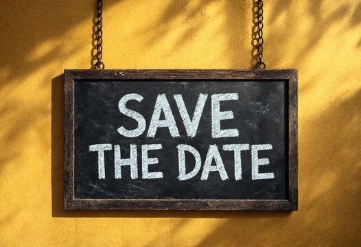 Black chalkboard sign with save the date written hanging on yellow wall background