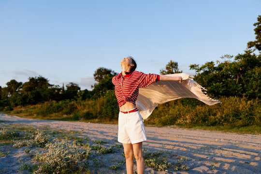 One woman wearing a striped red top and white shorts stands in a sunlit field, scarf flowing with the wind, expressing carefree summer fashion.
