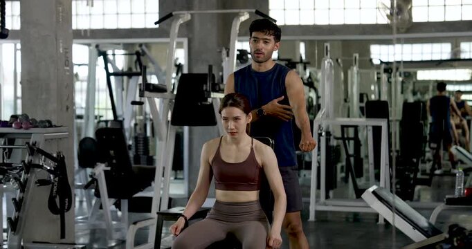 Young Asian couple exercising together in modern gym, man guiding woman during seated dumbbell workout, partner training for arm strength, encouragement, teamwork, wellness, relationship goals.