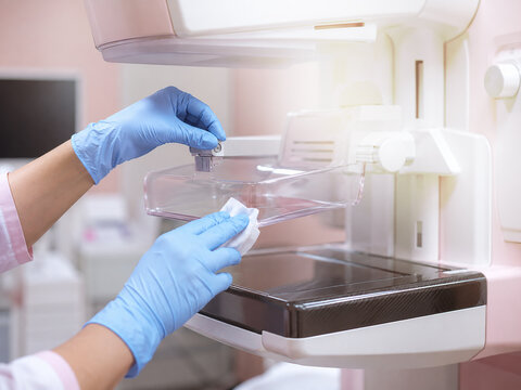 Close up of hands in blue nitrile gloves cleaning the clear compression paddle of a mammography machine with a disinfectant wipe. Generative AI.