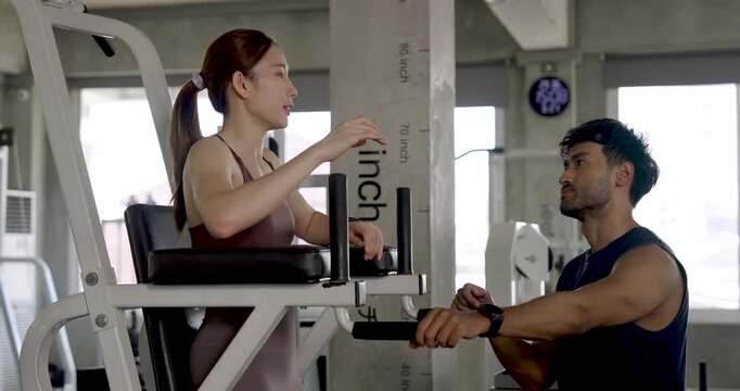 Young Asian couple training together in modern gym, woman uses knee raise station while man sits nearby guiding form, partner workout with support, leg strength, motivation, wellness routine.