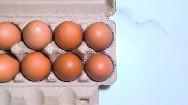 Hand opening a brown cardboard carton containing fresh organic brown eggs on a white marble surface.