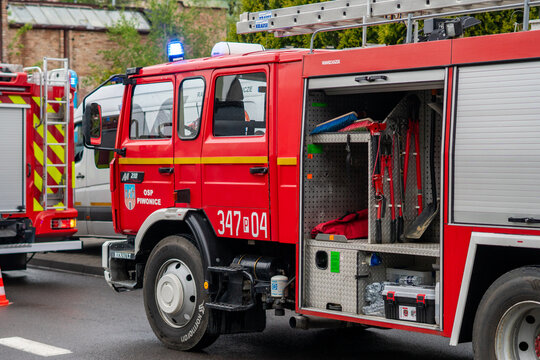 Red Renault fire engine of OSP Piwonice with open side equipment compartment standing on street with flashing blue lights during rescue operation.