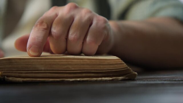 Woman reads book while sitting at table in dimly lit room, her finger tracing text on page close-up