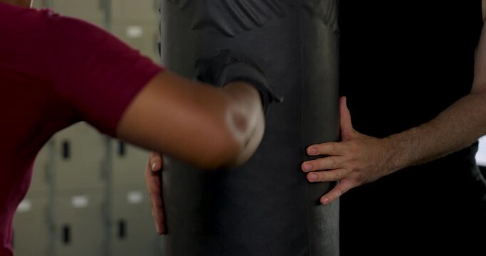 Close view of plus size African American woman punching heavy bag in modern gym while male trainer steadies bag and coaches technique, creating partner boxing workout for cardio, strength, endurance.