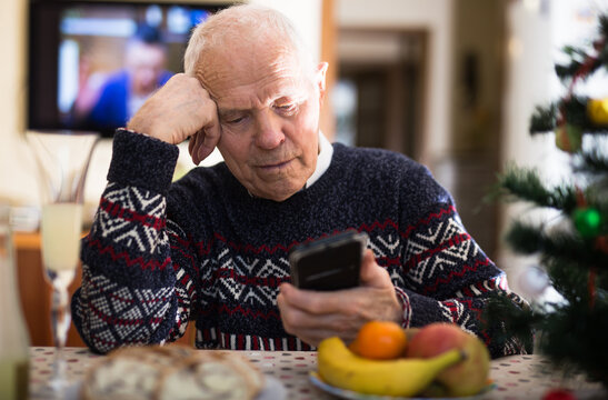 lonely senior man sitting at home table during Christmas holidays, looking at phone