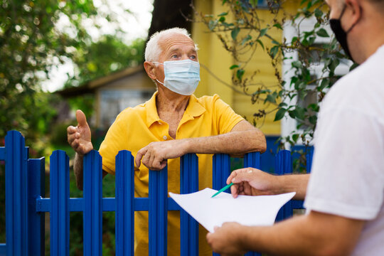 Grandfather in protective mask talking to census agent standing at fence of his country house