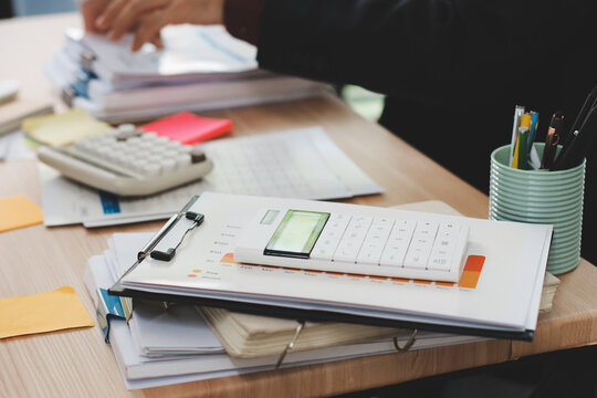 Accountant reviewing printed financial reports at messy office desk with calculator and sticky notes under soft window daylight for audit and corporate finance work