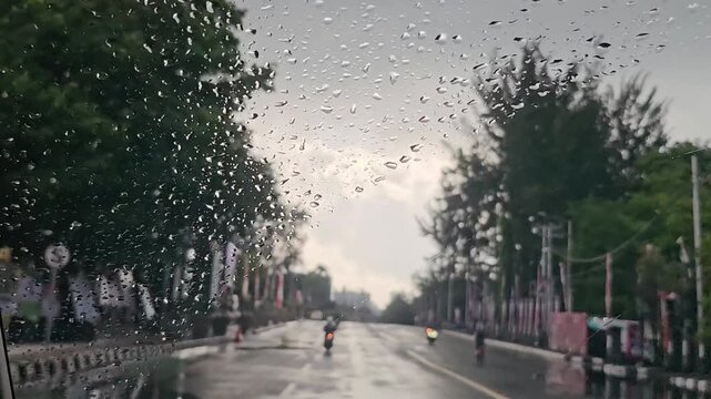 rain on the street,Raindrops streak across a car window, blurring the view ahead. Trees line the wet road, their leaves glistening. Lone motorcyclist rides through the downpour, unseen in the haze
