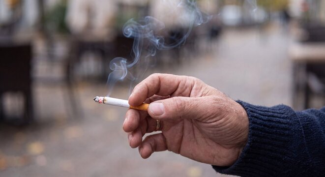 Middle aged hand holding lit cigarette emitting wispy smoke against blurred outdoor urban background setting with cigarette releases thin displays close person