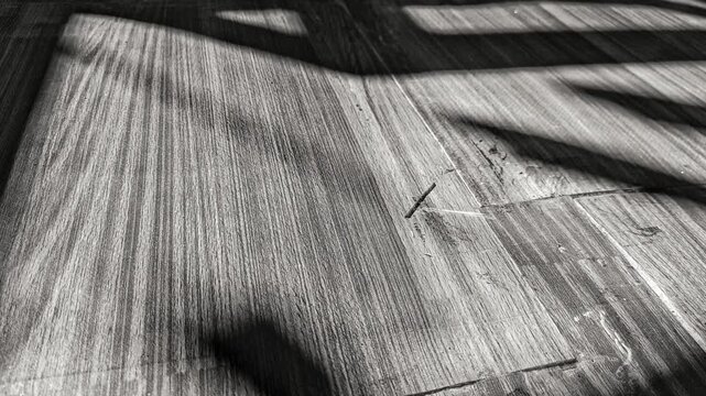Moody black and white footage of vertical moving shadow lines from a window casting over a rustic wooden floor texture, abstract cinematic background loop.