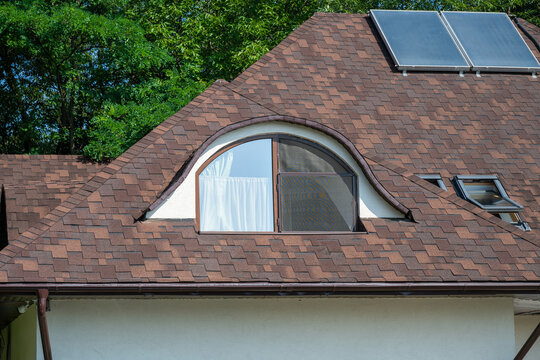 a detailed exterior view of a residential home's brown asphalt shingle roof, highlighting a prominent arched dormer window, modern skylights, and solar panels.