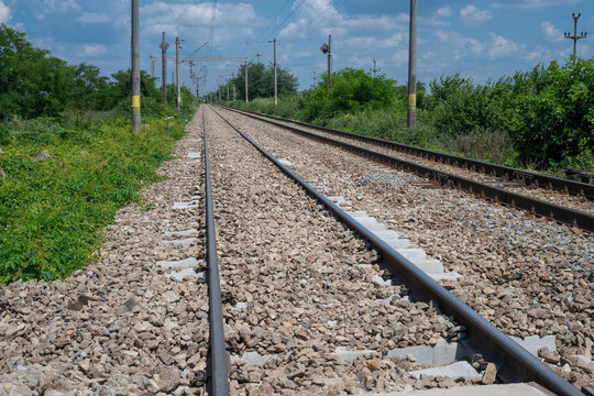 empty double railway tracks stretching toward the horizon, bordered by green vegetation and utility poles under a partly cloudy blue sky.