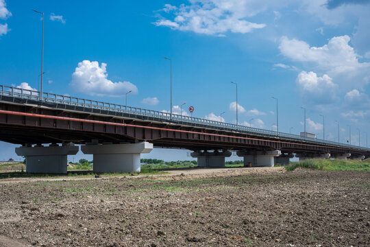 a long, sturdy highway bridge with concrete pillars and steel beams spanning across a tilled agricultural field under a bright blue sky with fluffy white clouds.