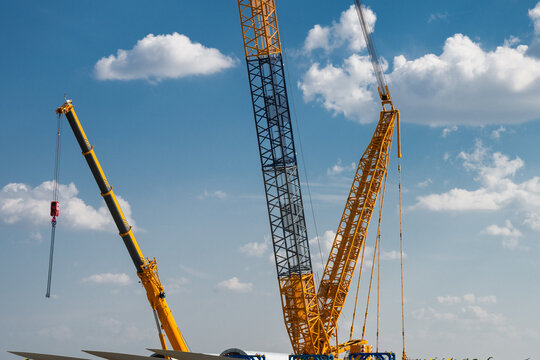 the towering yellow lattice and telescopic booms of heavy construction cranes reaching into a bright blue sky dotted with fluffy cumulus clouds.