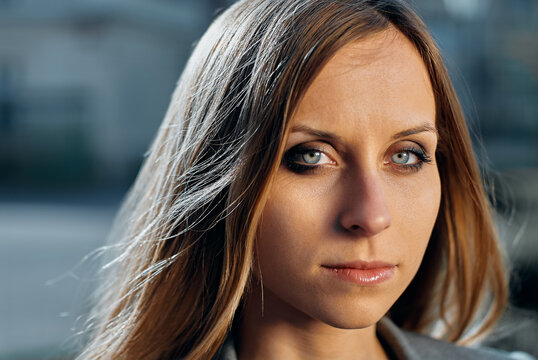 A close-up portrait of a serious young woman with striking blue eyes and long hair, photographed outdoors in natural light, conveying confidence, introspection and subtle professional elegance.