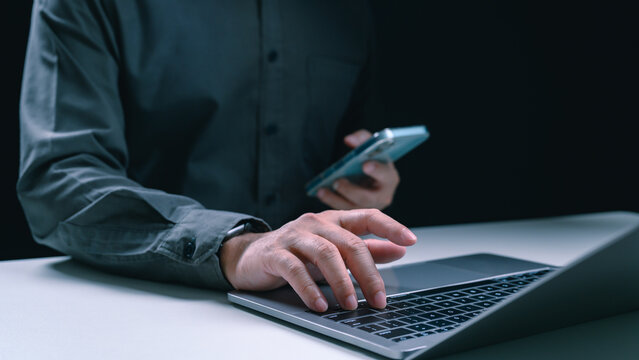 Man typing on a laptop while holding a smartphone in the dark