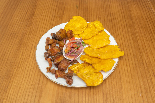 Plate of crispy fried pork, golden plantain chips, and fresh tomato and onion salsa on a wooden table.