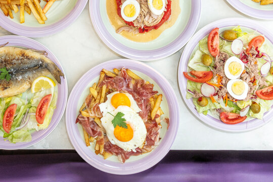 Overhead spread of huevos rotos with ham and fries, grilled sea bass, tuna salad, and Basque-style egg and pepper salad.