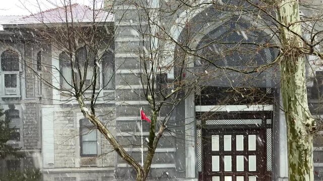 Static shot of the historic stone facade and ornate wooden entrance of an Istanbul building under a gentle snowfall with a fluttering Turkish flag in Turkey.
