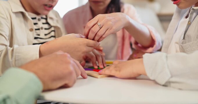 Family parents and children playing puzzle boardgame together. Hands assemble bright shapes at the table, cooperation, focus and motor learning at home. Concept of family bonding and play.